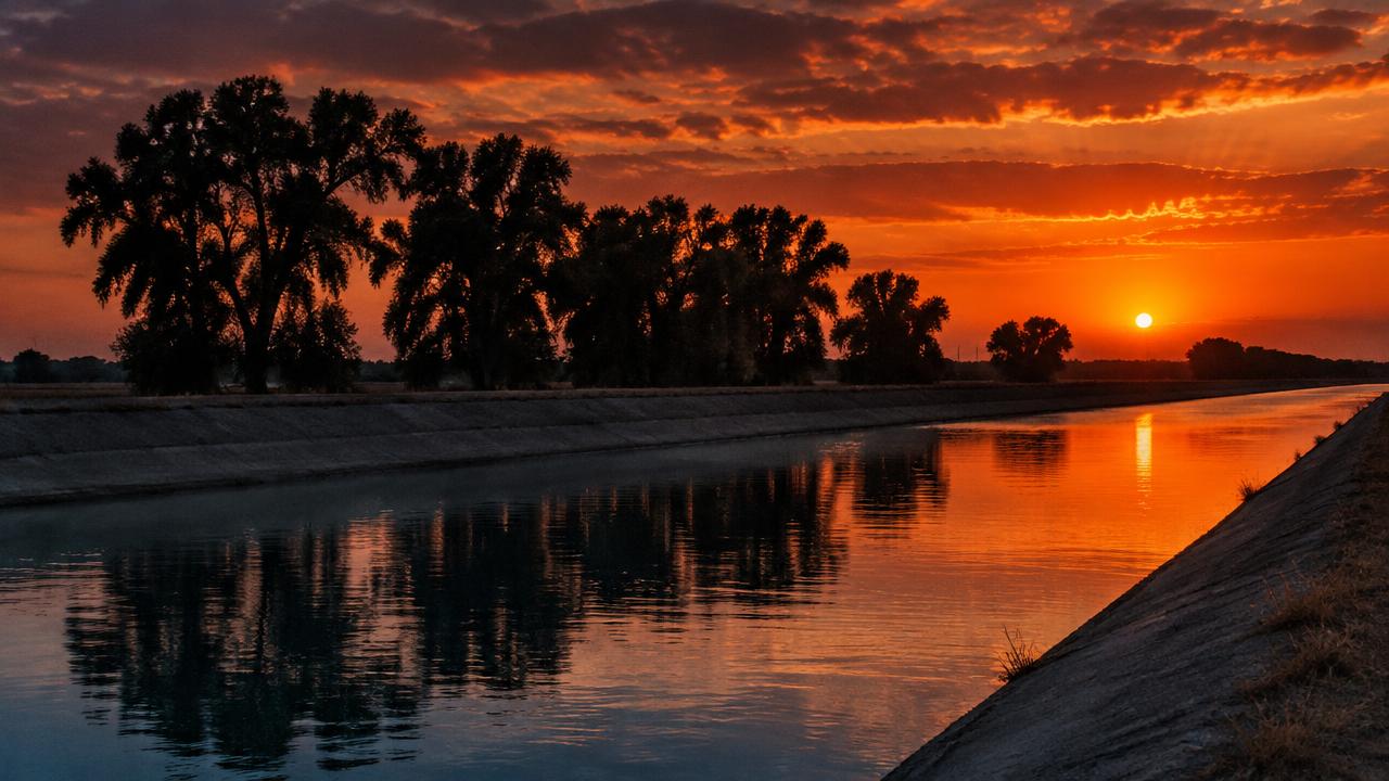 Irrigation canal at dusk under a burnt-orange sky, cottonwood trees reflected in the water.